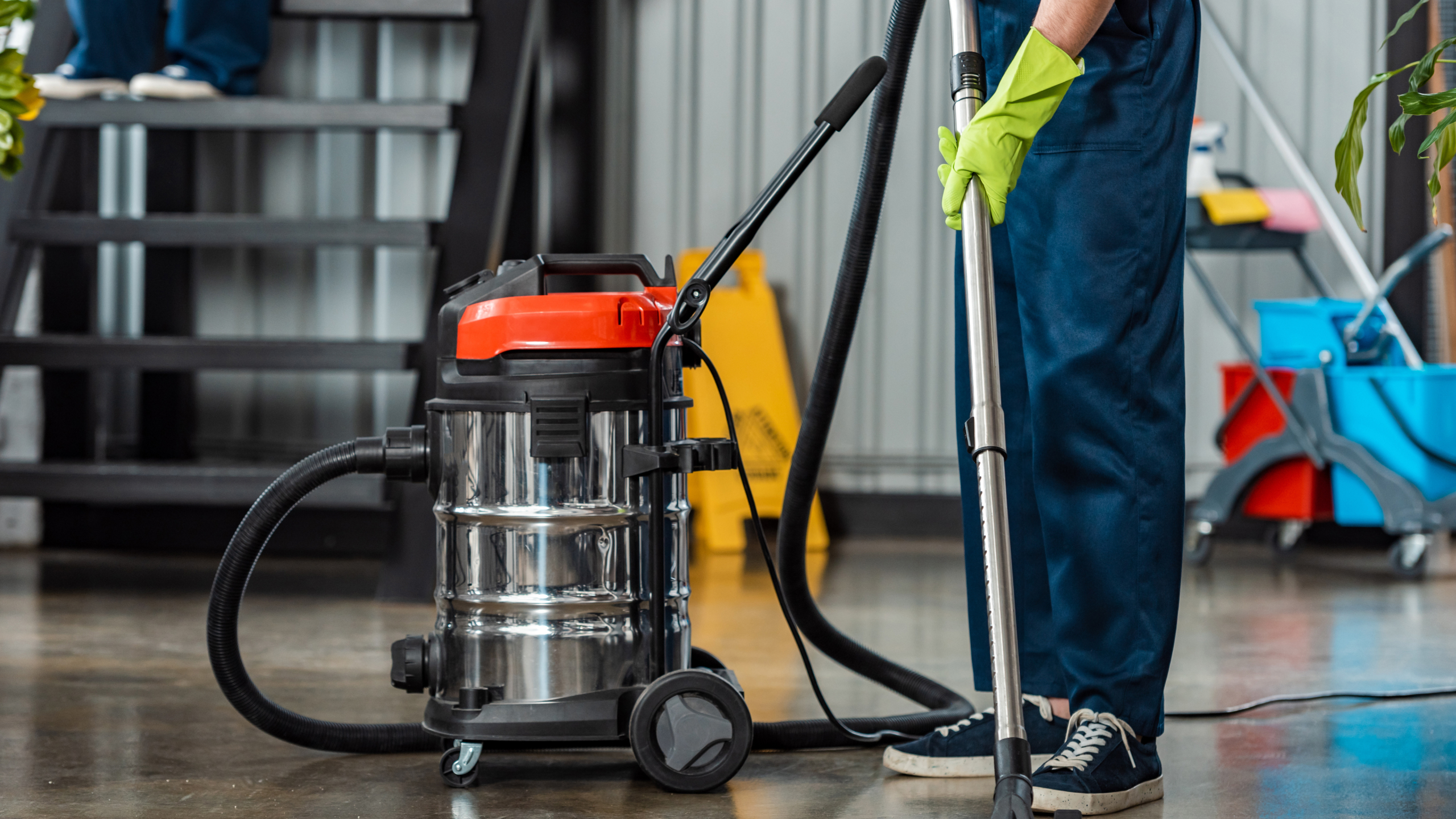 A person wearing green gloves is standing next to a wet/dry vacuum cleaner with a transparent tank, preparing to clean a concrete floor.