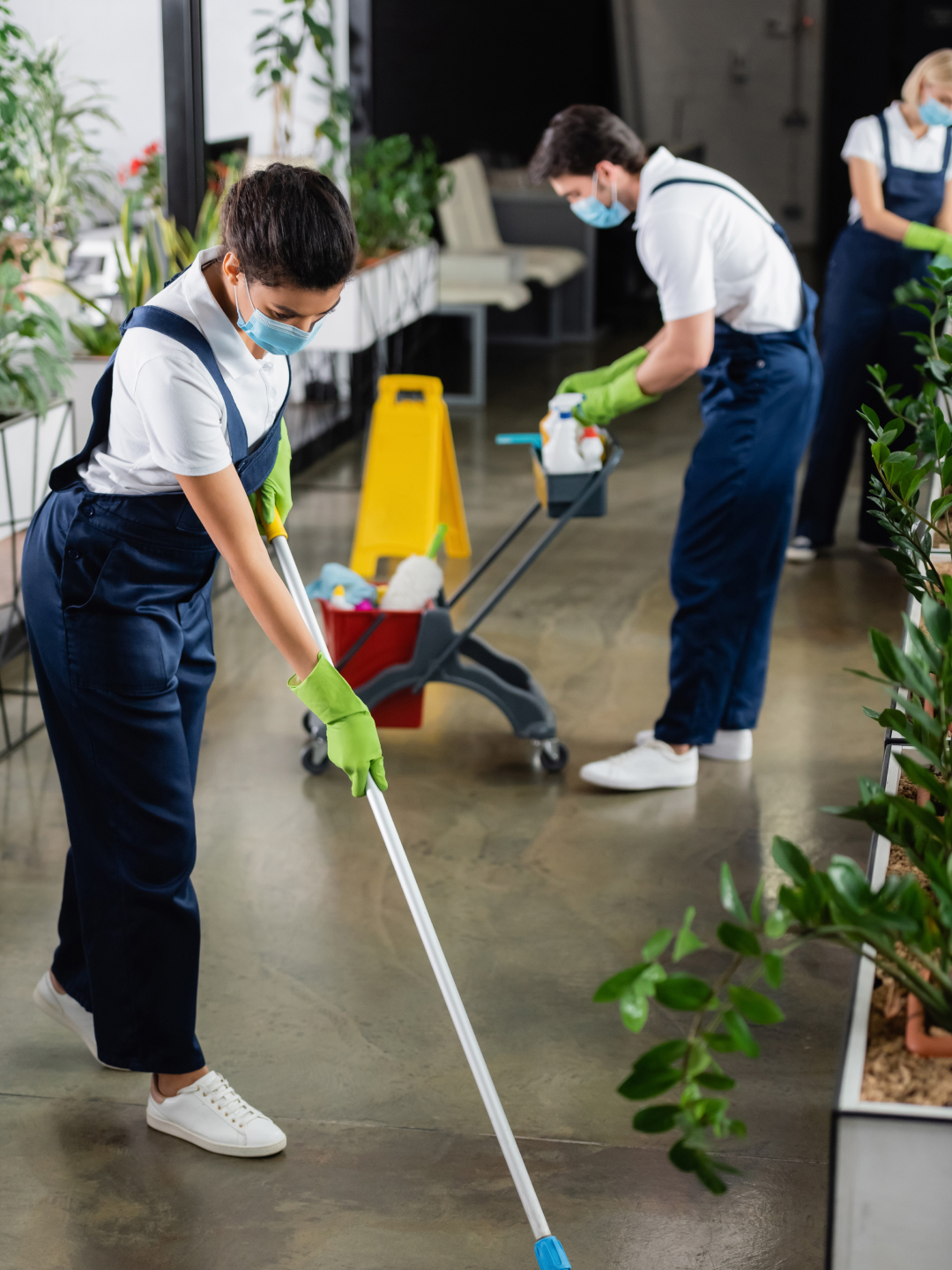 Two individuals wearing masks and gloves are cleaning indoors. One person is mopping the floor while the other is pushing a cart with cleaning supplies. Plants are visible in the background, and the space appears to be well-lit and modern.