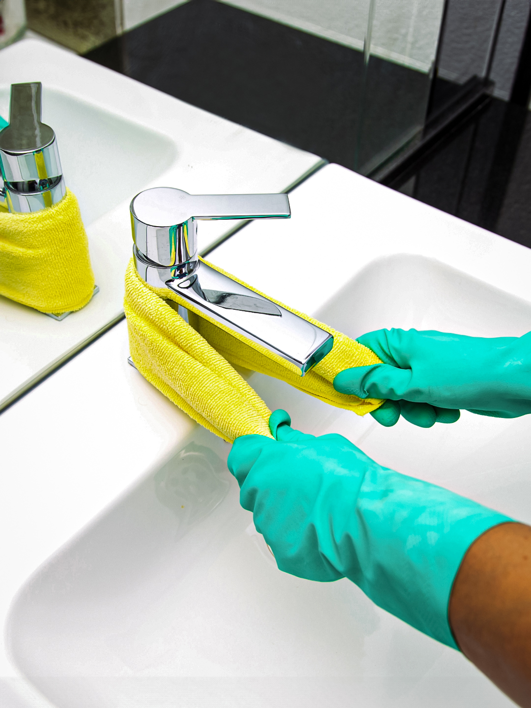 A person wearing green rubber gloves is cleaning a chrome faucet with a yellow cloth in a bathroom sink. The sink and faucet are shown in a bright, well-lit setting.